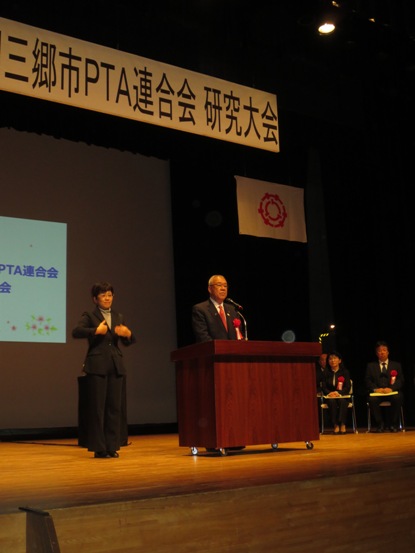 「三郷市PTA連合会研究大会」と書かれたボードが掛けられているステージ上で、スクリーンの前に設置された演台に立ち開会のあいさつの言葉を述べる三郷市長と、市長の横に立ち手話通訳をしている女性の写真