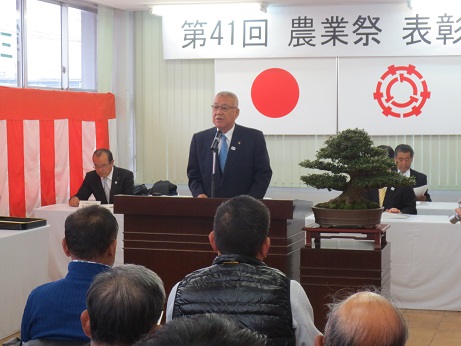 「第41回三郷市農業祭表彰式」と書かれた横断幕と国旗・三郷市旗が貼り付けられている壁の前で、立派な盆栽が飾られている花台と並ぶ演台に立ち挨拶の言葉を述べる三郷市長と、椅子に座って話を聞く関係者の写真