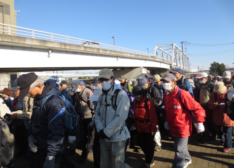 雲一つない青空の下、大勢の参加者と赤いジャンパーを着用したスタッフが同じ方向に向かって歩いている写真