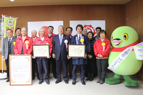 国旗と三郷市市旗が飾られた室内で市長を中央に右側に賞状を持ち法被を着た男性、左側に賞状を持ち赤色のジャケットを着た男性、他10名の関係者の方々とマスコットキャラクターかいちゃんが並んで立っている記念写真