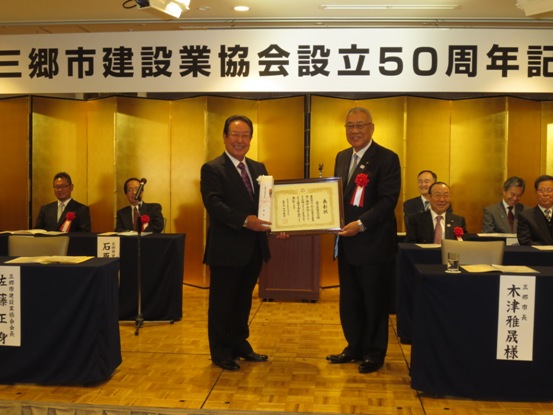 ステージに並んだ来賓席の中央で、記念品を右手に持った男性が、左手で三郷市長とともに表彰状を持っている記念写真