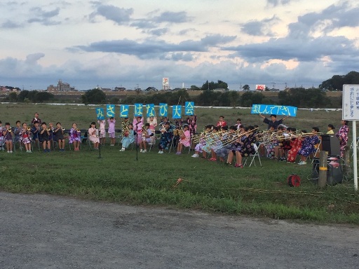 河川敷の広場に扇形に設置されたパイプ椅子に座った浴衣姿の児童達が演奏を行っている左後方で、一文字ずつ「みさとはなび大会」と書かれた大文字を一人ずつ持ち、右後方の男児が「楽しんで下さい」と書かれたボードを持ち上げている写真