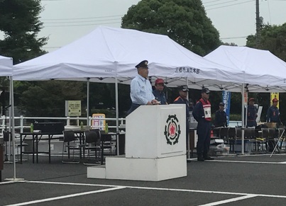 白い演台に立ちあいさつの言葉を述べる三郷市長と、白いテントの前に立って話を聞く消防団関係者の写真
