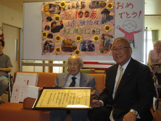 「祝100歳峠雅治様大正7年3月20日」の文字とたくさんの峠さんの写真と紙で作られたひまわりで囲んだ手作りのポスターが貼られ、「おめでとう」の文字と峠さんのイラストが描かれたホワイトボードを背に、オレンジ色のソファーに座った、峠雅治さんと三郷市長が表彰状を手に持って撮影した記念写真