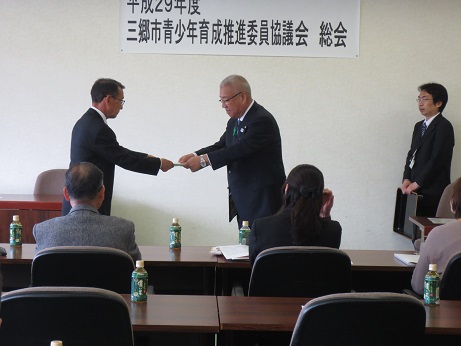 「平成29年度三郷市青少年育成推進委員協議会総会」とかかれたポスターが壁に貼ってあり、三郷市長が男性に文書を手渡し、ペットボトルのお茶が置かれたテーブルに座る参加者がその様子を見守っている写真