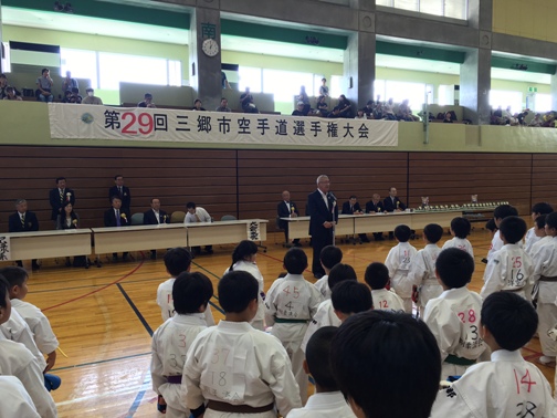 「第29回三郷市空手道選手権大会」と書かれた横断幕が2階席の壁に貼られた体育館内で、壁側に設置された長机の席に座っている関係者と道着を着た子供たちが向かい合って立ち、中央に設置されたマイクスタンドの前で挨拶を行っている市長の話を聞いている写真
