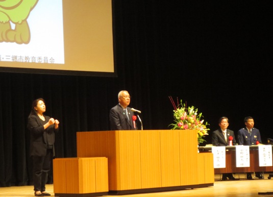 ステージ上のピンク・黄色・白の豪華な花々で彩られた花台と並ぶ演台に立ち挨拶の言葉を述べる三郷市長と手話通訳をする女性、ステージ上に配置された席に座り話を聞く胸章を付けた来賓者の写真
