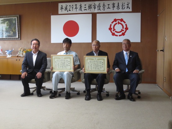 「平成29年度三郷市優秀工事表彰式」と書かれたポスターと国旗・三郷市旗が掛けられている壁の前で、受賞した2名の現場技術者が表彰状を両手で持ち、三郷市長と関係者の男性とともに記念撮影をしている写真