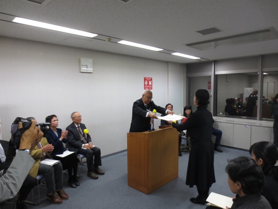 演台に立ち表彰状を手渡す三郷市長と受け取る黒い服を着用した女性、パイプ椅子に座り表彰の様子を見守る関係者の写真