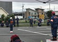 駐車場に立つ紺の制服と白いヘルメットを着用しオレンジ色のゼッケンをつけた消防団員と、その様子を見守る多くの消防隊員の写真