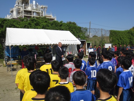 第34回三郷市長杯少年サッカー大会の開会式で、白いバザーテントの前に立ち話をする三郷市長と、整列し立って話を聞く黄色・グレー・青のユニフォームを着た子どもたちの写真