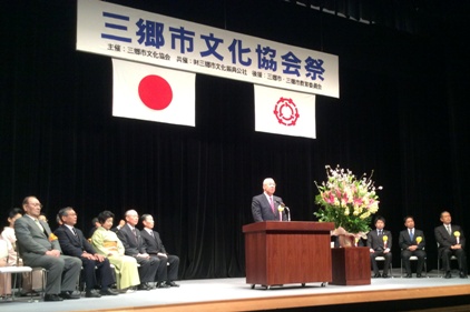 「三郷市文化協会祭」と書かれた横断幕や国旗と三郷市市旗が飾られたステージの檀上で挨拶を行っている市長の写真