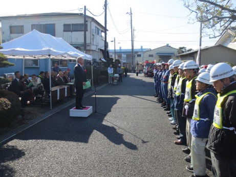 細い道路の右側にヘルメットを被った方々が横一列に並び、向かい合って小さな台の上に立っている市長が挨拶を行っている写真