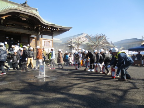玉蔵院の建物の前で、ヘルメットを被った参加者たちが「火」と書かれた的をめがけて水消火器を向けて放水している訓練の様子の写真