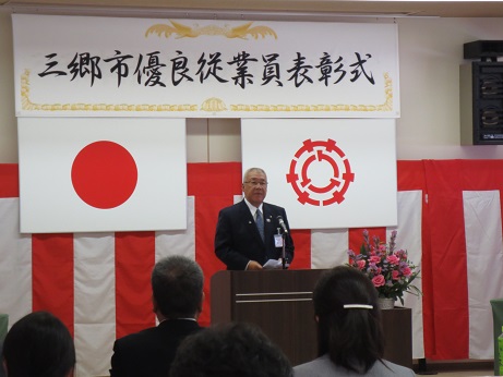 「三郷市優良従業員表彰式」と書かれた横断幕と国旗と三郷市旗、紅白幕が貼られた会場で、椅子に座っている受賞者と向かい合って演壇に立つ市長が挨拶を行っている様子の写真
