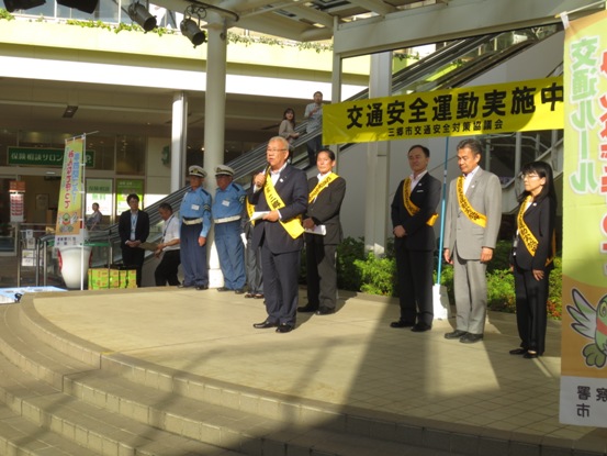 黄色地に「交通安全運動実施中」と書かれた横断幕の前で、キャンペーン活動前の挨拶の言葉を述べる黄色いタスキをかけた三郷市長と、市長の後ろに横一列に並んで話を聞いている同じタスキをかけた関係者たちの写真