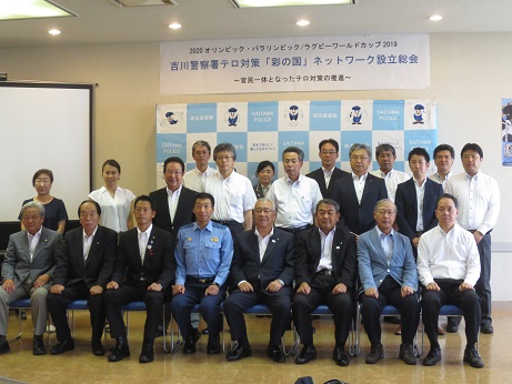 「吉川警察署テロ対策（彩の国）ネットワーク設立総会」と書かれた横断幕が壁に貼り付けられてあり、埼玉警察署のバックボード前に三郷市長と構成団体のメンバーが並び記念撮影をしている写真
