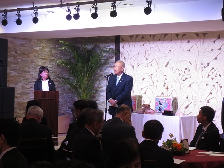 2つの酒樽が置かれた台の前であいさつの言葉を述べる三郷市長と、司会台に立つ女性、円形テーブルを囲んで座り話を聞く参加者の写真
