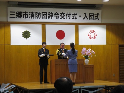 「三郷市消防団辞令交付式・入団式」と書かれた横断幕と国旗と三郷市旗などが壁に飾られた室内の演壇を挟んで、制服を着用した男性とスカートスーツを着た女性が向かい合って立ち辞令交付を行っている様子の写真