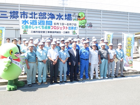「水道週間6月1日～6月7日公園のじゃぐち点検プロジェクト出陣式！」と書かれた横断幕が固定されているフェンスの前で、三郷市長と三郷市指定管工事業協同組合のメンバーが、タスキをかけたかいちゃんと「通水50周年」と書かれたのぼり旗とともに並んで記念撮影をしている写真