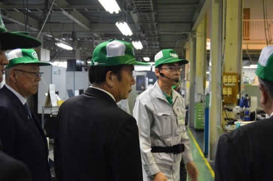 工場の中で、緑の帽子とグレーの作業服を着用し、マイクを付けた株式会社パーツ精工の社員が、上田清司埼玉県知事と三郷市長、その他訪問者に説明を行っている様子の写真