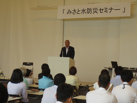 「みさと水防災セミナー」と書かれたポスターが天井から吊り下げてあり、白い演壇に立ち開会の挨拶をする三郷市長と席に座って話を聞く参加者の写真