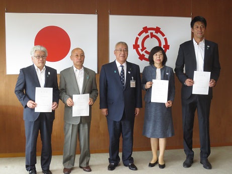 国旗と三郷市旗が吊り下げられている壁の前で、任命書を手に持った山本隆弘さん、坪井裕子さん、小阪秀史さん、田中彰則さんが市長を中心にして並んで立っている記念写真