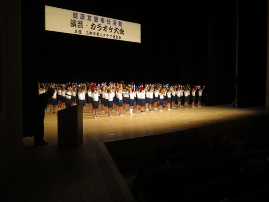 ステージ上には「演芸・カラオケ大会」と書かれた横断幕が吊るされており、キラキラと輝く色とりどりのポンポンを片手に持った大勢の丹後小の子どもたちが整列し両手を挙げている写真