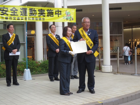 黄色地に「交通安全運動実施中」と書かれた横断幕の前で、黄色いタスキをかけた三郷市長と小林那智子会長が知事からのメッセージを手に持ち記念撮影をしている様子の写真