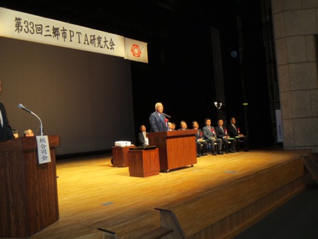 「第33回三郷市PTA研究大会」と書かれた横断幕が飾られた檀上で挨拶を行っている市長の写真