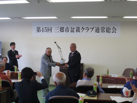 「第45回三郷市盆栽クラブ通常総会」と書かれたポスターが壁に貼ってあり、三郷市長にさつきの苗木を手渡しているグレーのスーツを着た男性と、ペットボトルのお茶が置かれた席に座り拍手をする参加者の写真