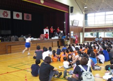 ステージの上で抽選を行っている赤色のシャツを着た4名のスタッフが立ち、左側から女の子が走ってスタッフに駆け寄っている抽選会の様子の写真