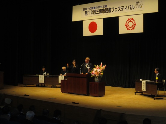 「日本一の読書のまち三郷 第12回三郷市読書フェスティバル」の吊り看板が設置された舞台上で、演壇に立って話をしている市長の写真