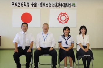 平成26年度全国・関東大会出場選手激励会と書かれた横断幕、国旗と三郷市旗が飾られた室内で水泳で優秀な成績を収めた南中学校の佐藤果歩さんと男性と女性が市長を挟んで横一列に並んで椅子に座っている記念写真