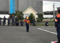 駐車場に立って敬礼をするオレンジと紺の制服と黄色いヘルメットを着用した少年消防クラブの男の子と、その様子を見守る消防団員の写真