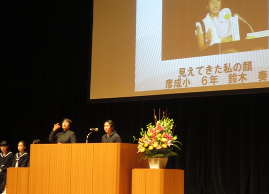 ステージ上のピンク・黄色・白の豪華な花々で彩られた花台と並ぶ演台に立ち、背後のスクリーンに映し出されている「見えてきた私の顔」のテーマについて発表をする鈴木奏さんと、隣で手話通訳をしている女性の写真