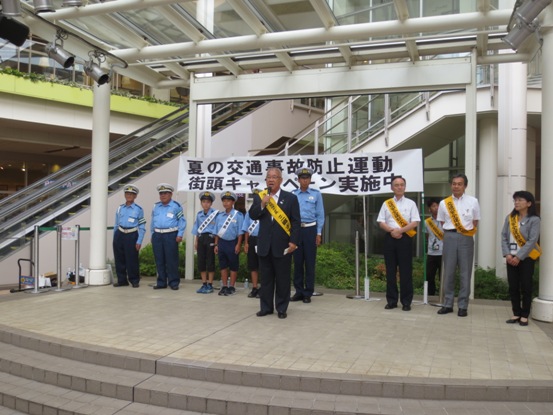 「夏の交通事故防止運動街頭キャンペーン実施中」と書かれた横断幕の前で、キャンペーン前に挨拶の言葉を述べる黄色のたすきをかけた三郷市長と、市長の後ろで背筋を伸ばして立つ一日警察署長の吉川市、松伏町、三郷市の子どもたちと関係者の写真
