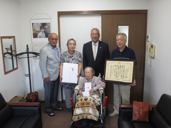 三郷市長が、記念品を手に持ち車椅子に座る佐藤知江子さんと表彰状と記念品を手に持つご家族とともに記念撮影をしている写真