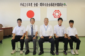 平成26年度全国・関東大会出場選手激励会と書かれた横断幕、国旗と三郷市旗が飾られた室内で空手で優秀な成績を収めた栄中学校の3名の男子生徒と男性教師が市長を挟んで横一列に並んで椅子に座っている記念写真