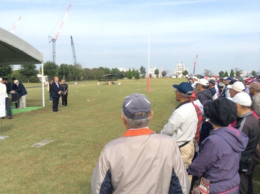 グリーンの芝生の上でグラウンドゴルフ大会の参加者と向かい合って立っている市長が挨拶を行っている様子の写真