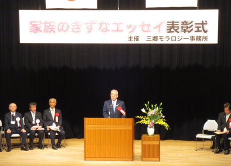 「家族のきずなエッセイ表彰式」と書かれた横断幕がステージ上に吊るしてあり、ピンク・黄色・白の豪華な花々で彩られた花台と並ぶ演台に立ち挨拶の言葉を述べる三郷市長と、ステージ上に置かれたパイプ椅子に座り話を聞く胸章を付けた来賓者の写真