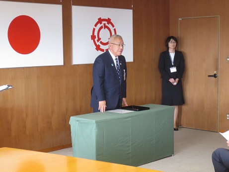 国旗と三郷市旗が吊り下げられている壁の前で、緑のカバーが掛けられた台の後ろに立ち話をする三郷市長の写真