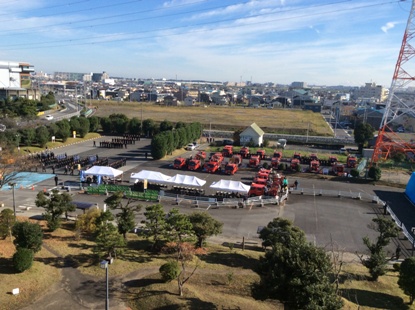 手前に4つのテント、右側に沢山の消防車が停まり、左側に大勢の消防団員が整列している様子を高台から撮影した写真