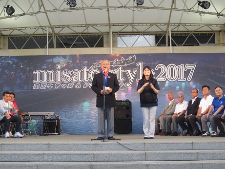 手前にスピーカーや電子ピアノが置かれた「misatostyle2017」と書かれたバックパネルを背にステージに立ってあいさつの言葉を述べる三郷市長と市長の横で手話通訳をしている女性、ステージに並んだパイプ椅子に座り話を聞く関係者たちの写真