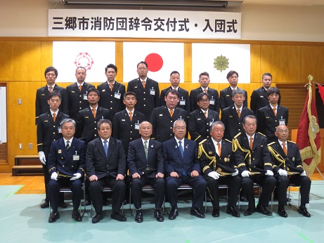 三郷市旗・国旗・消防旗の上に「三郷市消防団辞令交付式・入団式」と書かれた横断幕が吊り下げられた壁の前で、三郷市長と制服を着用した消防団のメンバーが並んで記念撮影をしている写真