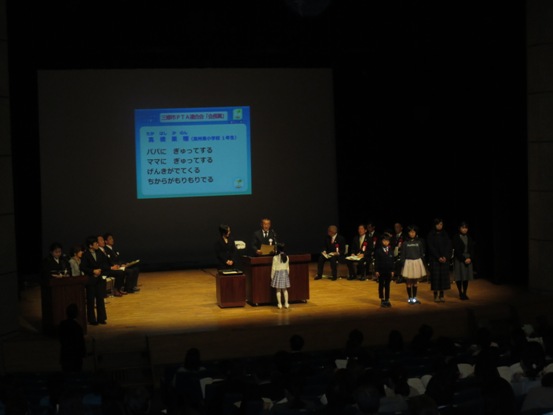 三行詩である「パパにぎゅってするママにぎゅってするげんきがでてくるちからがもりもりでる」が映し出されたスクリーンの前で、演台に立ち表彰状を読み上げる三郷市長と演台の前で内容を聞く女の子の写真