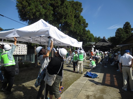 白いヘルメットと防犯パトロールベストを着用した参加者が、青空の下、協力し合いながら白いテントを設営している訓練の様子の写真
