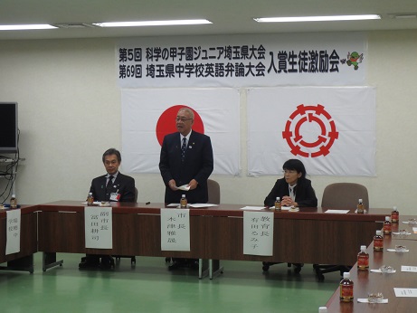 「第5回科学の甲子園ジュニア埼玉県大会第69回埼玉県中学校英語弁論大会入賞生徒激励会」と書かれた横断幕と国旗・三郷市旗が貼り付けられた壁の前で、コの字型に並んだ長机の席を立ち、挨拶の言葉を述べる三郷市長と、席札が貼られペットボトルのお茶が置かれている、市長と横並びの席に座り話を聞く副市長と教育長の写真