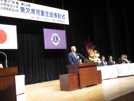 「第38回無欠席児童生徒表彰式」と書かれた横断幕が飾られたステージの演壇で挨拶を行っている市長の写真