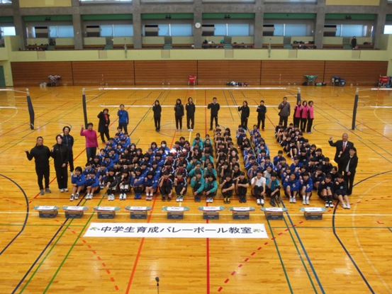 「中学校育成バレーボール教室」と書かれたポスターの後ろに各学校ごとに整列し体育座りをする中学生と、三郷市長、元日本代表バレーボール選手、関係者がバレーボールネットの前に集まり2階部分から記念撮影をしている写真